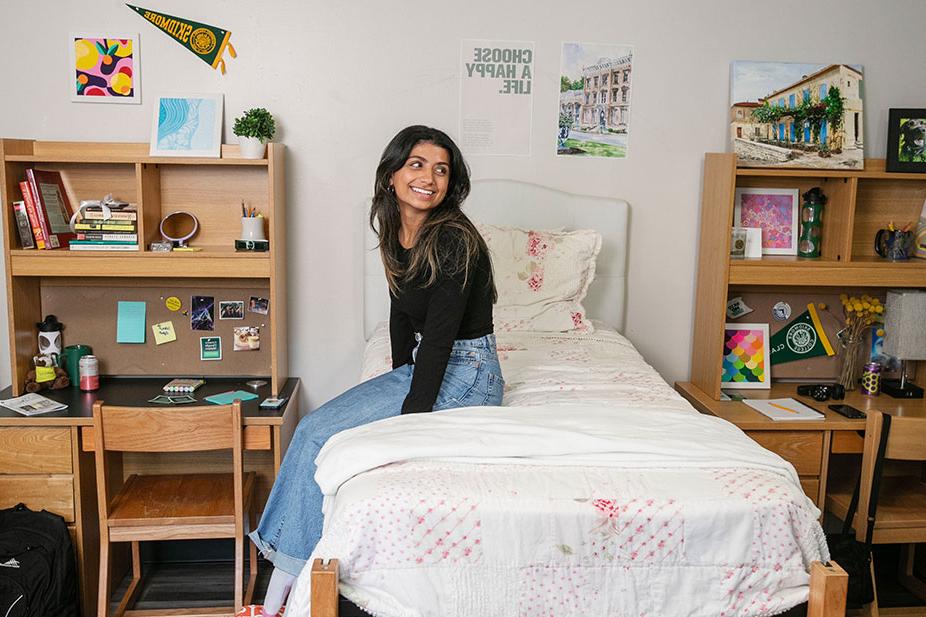 college-age woman smiles while sitting on her bed in a decorated dorm room that features bookshelves and colorful decorations